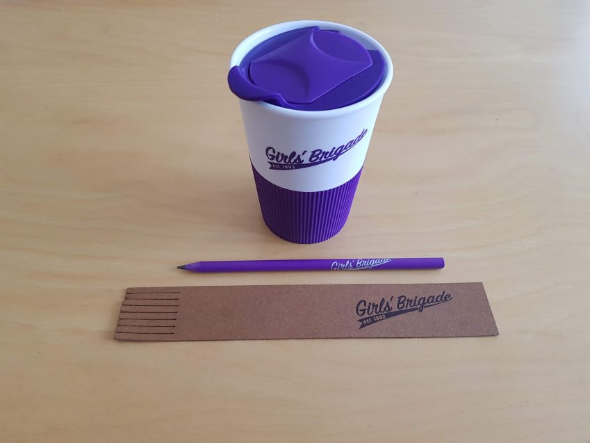 Girls' Brigade Trading products, a purple and white travel mug, a purple pencil and a brown bookmark on a table